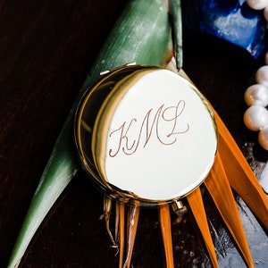 May include: A gold compact mirror with the initials "K.M.L." engraved on the lid. The mirror is surrounded by a green leaf and an orange flower.