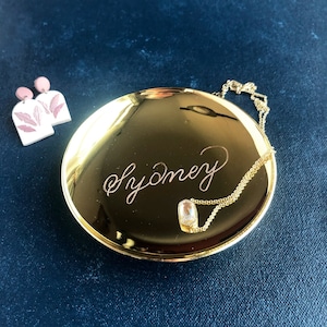May include: A gold-colored trinket dish with the word "Sydney" inscribed on the surface. A gold chain necklace with a clear stone pendant is resting on the dish.