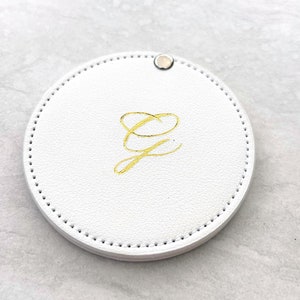 Może przedstawiać: Okrągłe, białe, skórzane lusterko kompaktowe z złotym monogramem 'G'.