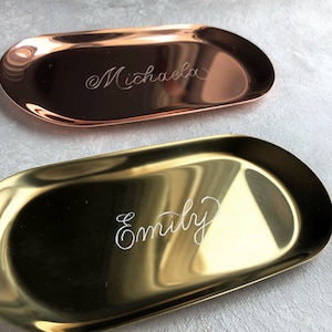 Könnte beinhalten: Zwei ovale Metallschalen, eine goldfarben und eine kupferfarben, mit den eingravierten Namen "Emily" und "Michaela".
