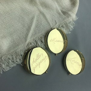 May include: Three gold-colored compact mirrors with engraved text. The first mirror says "hello gorgeous", the second says "Mirana", and the third says "Leona".