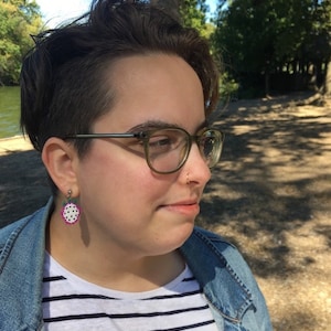 May include: A person with short, dark hair wearing glasses and a denim jacket over a striped shirt. They have a small, gold nose ring and a watermelon-shaped earring.