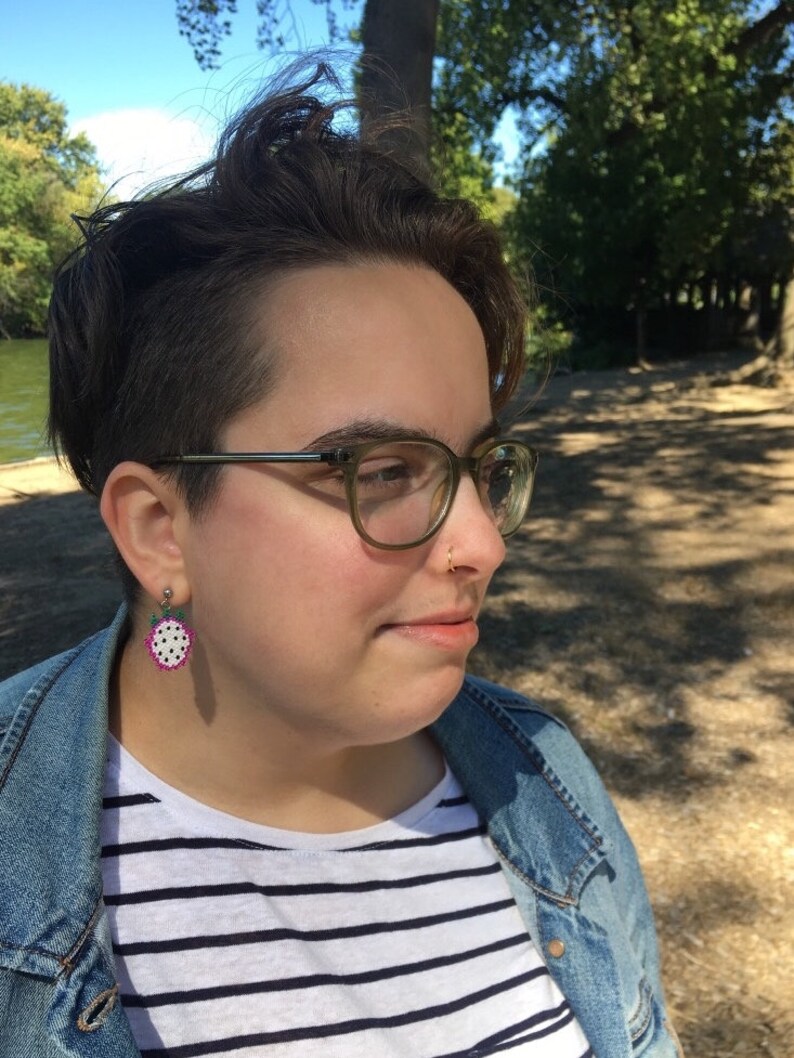 May include: A person with short, dark hair wearing glasses and a denim jacket over a striped shirt. They have a small, gold nose ring and a watermelon-shaped earring.