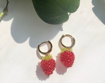 May include: A pair of gold hoop earrings with beaded strawberry charms. The strawberries are red with green beaded tops. The earrings are displayed on a white surface with a green leaf in the background.