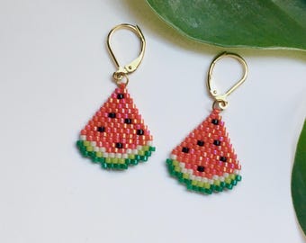 May include: A pair of gold-colored hoop earrings, each featuring a beaded watermelon slice design. The earrings have a red interior with black seed details, a green rind, and a gold-colored clasp. The background is white with a green leaf.