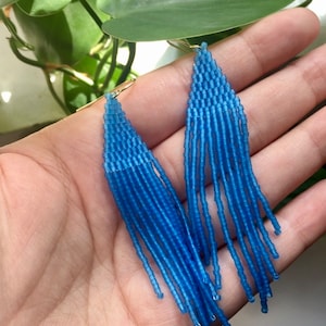 May include: A pair of long, blue beaded earrings with a triangular shape at the top. The earrings are made of small, round beads that are strung together to create a fringe effect.