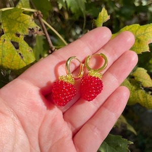 May include: A pair of gold hoop earrings with red beaded strawberry charms. The earrings are held in a hand against a green leafy background.