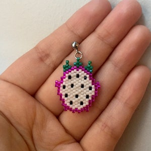 May include: A white and pink beaded dragon fruit earring with green leaves. The earring is on a silver hook.