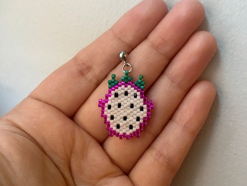 May include: A white and pink beaded dragon fruit earring with green leaves. The earring is on a silver hook.