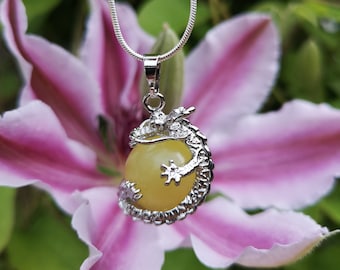 May include: A silver necklace with a pendant featuring a yellow orb embraced by a dragon design. The pendant hangs from a delicate silver chain. The background is a blurred image of a pink and white flower.
