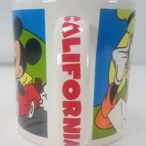 Puede incluir: Taza de cer&aacute;mica blanca con la inscripci&oacute;n "CALIFORNIA" en rojo en el asa. La taza presenta im&aacute;genes de dibujos animados de Mickey Mouse conduciendo un coche rojo, Donald Duck y Goofy.