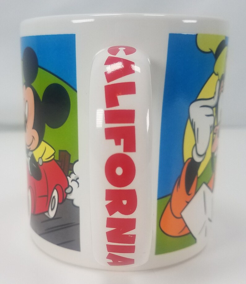 Puede incluir: Taza de cer&aacute;mica blanca con la inscripci&oacute;n "CALIFORNIA" en rojo en el asa. La taza presenta im&aacute;genes de dibujos animados de Mickey Mouse conduciendo un coche rojo, Donald Duck y Goofy.