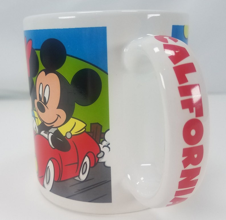Puede incluir: Taza de cer&aacute;mica blanca con una imagen de dibujos animados de Mickey Mouse y Minnie Mouse conduciendo un coche rojo. El asa de la taza tiene la palabra "CALIFORNIA" impresa en rojo.