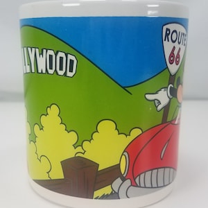 Puede incluir: Una taza de cer&aacute;mica blanca con una ilustraci&oacute;n colorida de Mickey Mouse conduciendo un coche rojo con un cartel de la Ruta 66. El fondo presenta una colina verde con la palabra "HOLLYWOOD" en letras blancas.