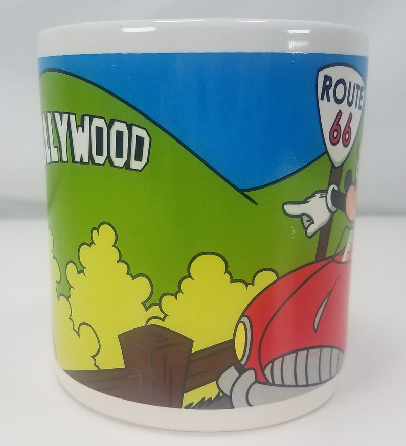 Puede incluir: Una taza de cer&aacute;mica blanca con una ilustraci&oacute;n colorida de Mickey Mouse conduciendo un coche rojo con un cartel de la Ruta 66. El fondo presenta una colina verde con la palabra "HOLLYWOOD" en letras blancas.