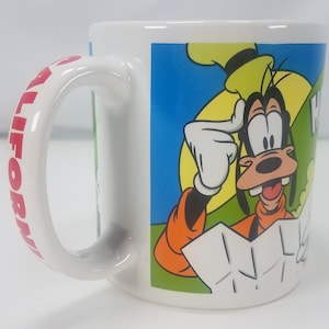 Puede incluir: Taza de cer&aacute;mica blanca con la inscripci&oacute;n "California" en rojo en el asa. La parte delantera presenta una imagen de dibujos animados de Goofy con una expresi&oacute;n confusa, con un sombrero amarillo y una camisa naranja, sosteniendo un mapa.