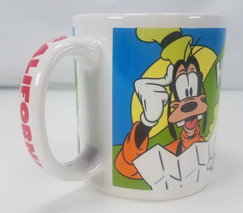 Puede incluir: Taza de cer&aacute;mica blanca con la inscripci&oacute;n "California" en rojo en el asa. La parte delantera presenta una imagen de dibujos animados de Goofy con una expresi&oacute;n confusa, con un sombrero amarillo y una camisa naranja, sosteniendo un mapa.