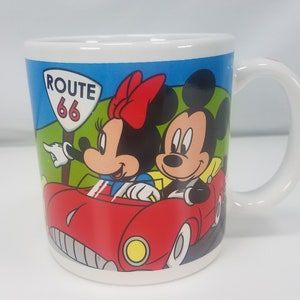 Puede incluir: Una taza de cer&aacute;mica blanca con una ilustraci&oacute;n colorida de Mickey Mouse y Minnie Mouse conduciendo un coche rojo por la Ruta 66.