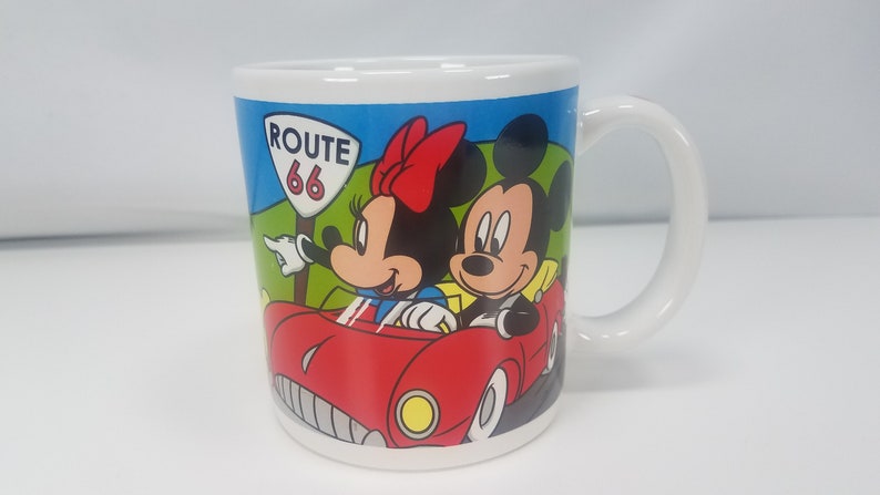 Puede incluir: Una taza de cer&aacute;mica blanca con una ilustraci&oacute;n colorida de Mickey Mouse y Minnie Mouse conduciendo un coche rojo por la Ruta 66.
