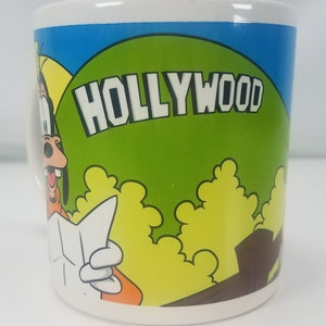 Puede incluir: Una taza de cer&aacute;mica blanca con una imagen de dibujos animados de Goofy sosteniendo un mapa. La taza tiene un fondo azul y verde con la palabra "HOLLYWOOD" en letras blancas.