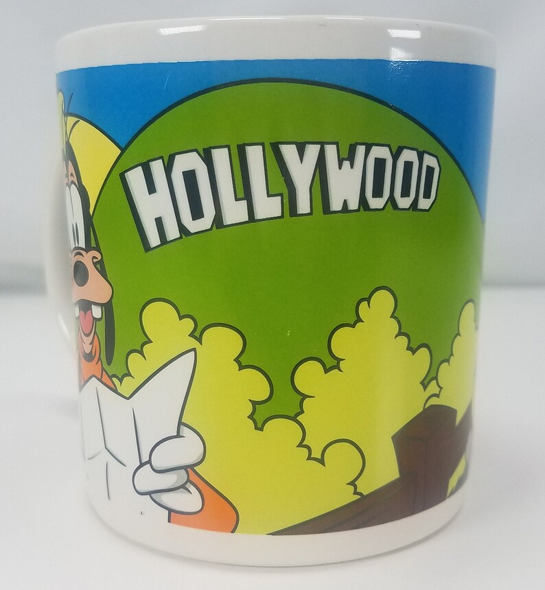Puede incluir: Una taza de cer&aacute;mica blanca con una imagen de dibujos animados de Goofy sosteniendo un mapa. La taza tiene un fondo azul y verde con la palabra "HOLLYWOOD" en letras blancas.