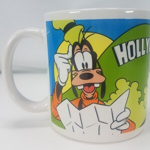 Puede incluir: Taza de cer&aacute;mica blanca con una imagen de dibujos animados de Goofy mirando un mapa con el texto "HOLLYWOOD" en el lateral.