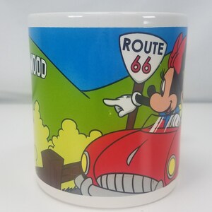 Puede incluir: Una taza de cer&aacute;mica blanca con una ilustraci&oacute;n colorida de Minnie Mouse conduciendo un coche rojo por la Ruta 66. El fondo presenta una colina verde con la palabra "HOLLYWOOD" parcialmente visible.