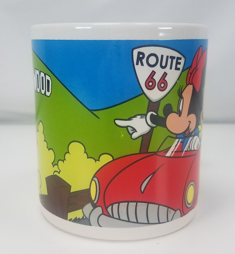 Puede incluir: Una taza de cer&aacute;mica blanca con una ilustraci&oacute;n colorida de Minnie Mouse conduciendo un coche rojo por la Ruta 66. El fondo presenta una colina verde con la palabra "HOLLYWOOD" parcialmente visible.