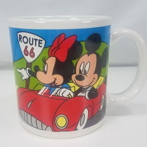 Puede incluir: Taza de cer&aacute;mica blanca con una ilustraci&oacute;n colorida de Mickey Mouse y Minnie Mouse conduciendo un coche rojo por la Ruta 66. La taza tiene un asa negra.