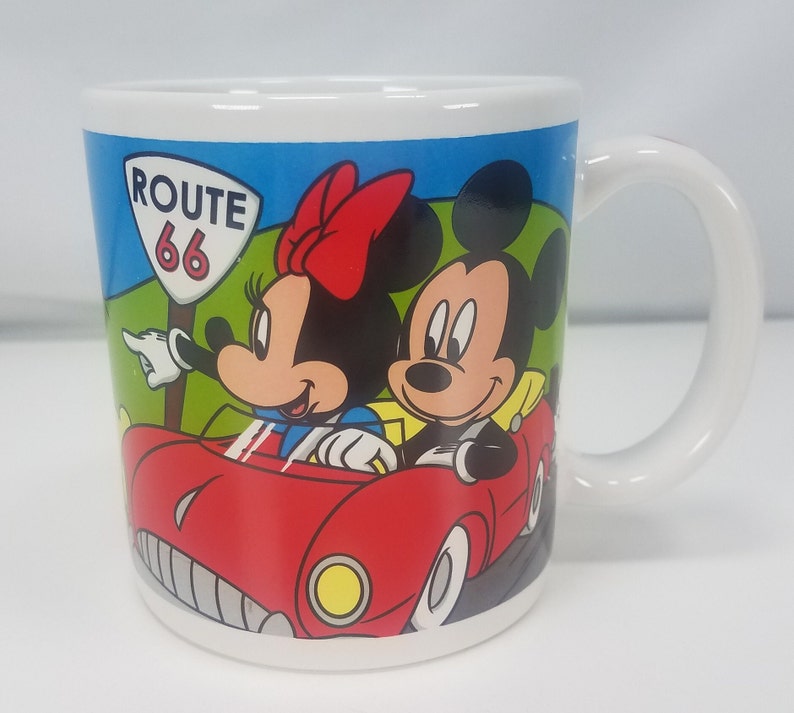 Puede incluir: Taza de cer&aacute;mica blanca con una ilustraci&oacute;n colorida de Mickey Mouse y Minnie Mouse conduciendo un coche rojo por la Ruta 66. La taza tiene un asa negra.