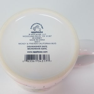 Puede incluir: Taza de cer&aacute;mica blanca con asa rosa. La taza presenta un dise&ntilde;o de Mickey Mouse y sus amigos. La parte inferior de la taza tiene una etiqueta con el texto "Everyone loves to get applause" y el logotipo e informaci&oacute;n de la empresa. La etiqueta tambi&eacute;n incluye el texto "Dishwasher Safe. Microwave Safe. www.applause.com" y un c&oacute;digo de barras.
