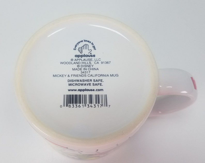 Puede incluir: Taza de cer&aacute;mica blanca con asa rosa. La taza presenta un dise&ntilde;o de Mickey Mouse y sus amigos. La parte inferior de la taza tiene una etiqueta con el texto "Everyone loves to get applause" y el logotipo e informaci&oacute;n de la empresa. La etiqueta tambi&eacute;n incluye el texto "Dishwasher Safe. Microwave Safe. www.applause.com" y un c&oacute;digo de barras.