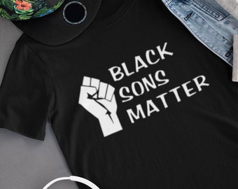 May include: Black t-shirt with a white raised fist and the text "Black Sons Matter". The shirt is laid flat on a gray surface with a green and black floral patterned hat and white headphones.