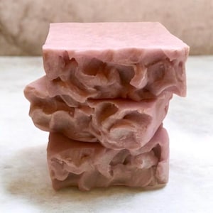 May include: Three stacked bars of handmade soap. The soap is a light pink color with a textured, uneven surface. The edges of the soap have a rough, organic appearance. The soap is stacked on top of each other.