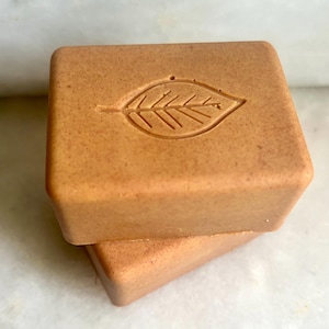 May include: Two square, light brown soap bars stacked on top of each other. The top bar has a leaf design carved into it. The soap has a natural, organic appearance.
