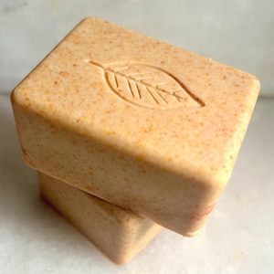 May include: Two rectangular bars of peach-colored soap stacked on top of each other. The top bar features a debossed leaf design on its surface. The soap has a textured, speckled appearance with small orange and brown particles throughout the material.