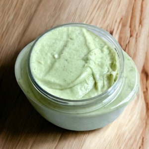 May include: A close-up of a green, creamy substance in a clear glass jar. The jar is on a wooden surface.