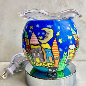 May include: A blue and white electric oil warmer with a whimsical village scene. The spherical warmer features a night sky design with stars, a crescent moon, and colorful buildings. It has a clear glass dish and sits on a silver base.
