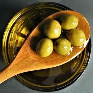 Puede incluir: Una cuchara de madera llena de aceitunas verdes en aceite de oliva. Las aceitunas est&aacute;n dispuestas en c&iacute;rculo alrededor de la cuchara.