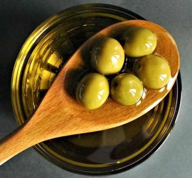 Puede incluir: Una cuchara de madera llena de aceitunas verdes en aceite de oliva. Las aceitunas est&aacute;n dispuestas en c&iacute;rculo alrededor de la cuchara.