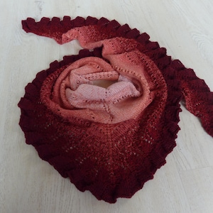 以下が含まれることがあります： 赤、ピンク、オレンジの色合いのクロシェのショール。繊細なレース模様で、縁はフリルになっています。