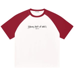 Può includere: Una t-shirt bianca a maniche corte raglan con maniche rosso scuro e scollo rotondo. Al centro del petto è presente la scritta nera "Wrong kind of witch" in un carattere in stile gotico, con un piccolo simbolo di linea orizzontale sotto.