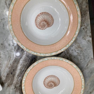 May include: Two pink and white ceramic soup bowls with a seashell design. The bowls have a scalloped edge and a gold trim.