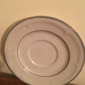 May include: A white ceramic saucer with a blue rim and a scalloped edge. The saucer is designed for a teacup or coffee cup.