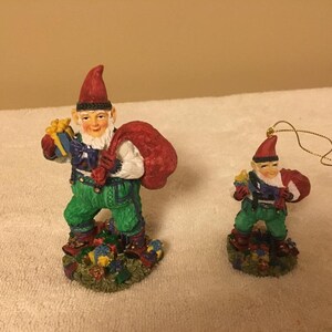 Puede incluir: Dos gnomos navideños, uno más grande que el otro, sostienen regalos y sacos. El gnomo más grande está de pie sobre una base con un diseño verde, marrón y azul. El gnomo más pequeño es un adorno con una cuerda dorada.