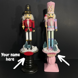 May include: Two custom nutcracker figurines, one in a traditional red and gold soldier uniform and the other in a pink and blue ice cream cone design. Both are standing on decorative bases with snow-covered trees.