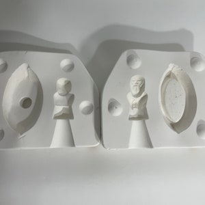 May include: Two white ceramic molds for making resin crafts. One mold has a figure of a person with a beard and a round object. The other mold has a figure of a person and a long oval shape.