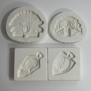 May include: Four white ceramic molds for making jewelry or other crafts. The molds feature Native American designs, including a chief's headdress, a feather, and an arrowhead.