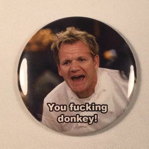 May include: Round button with a glossy finish featuring a portrait of a man with blonde hair and a white chef's coat. The text "You fucking donkey!" is printed in black on the button.
