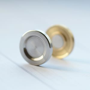 Könnte beinhalten: Zwei kreisförmige Metallobjekte mit einer vertieften Mitte. Ein Objekt ist silberfarben mit einer gebürsteten Oberfläche, während das zweite, leicht dahinter positionierte Objekt goldfarben mit einer glatten, reflektierenden Oberfläche ist.